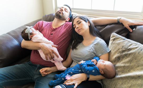 Tired Hispanic caring parents with newborn twins relaxing on sofa