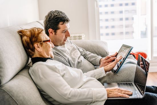 Side view of happy aged mother learning using laptop with help of adult son while sitting on sofa at home